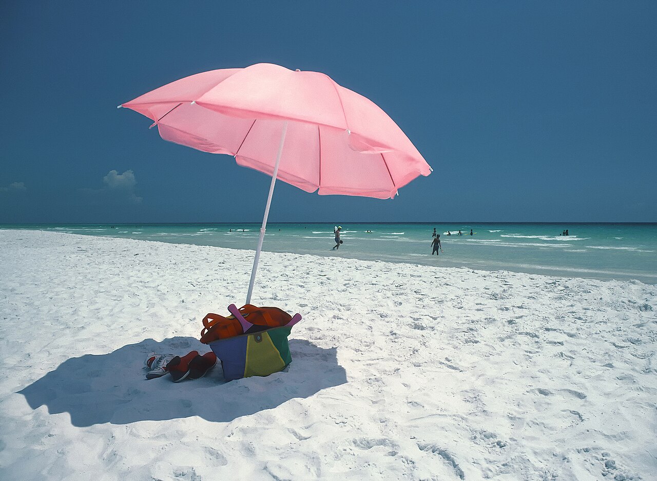 A pink beach umbrella on white sand beside emerald water in the Florida Panhandle